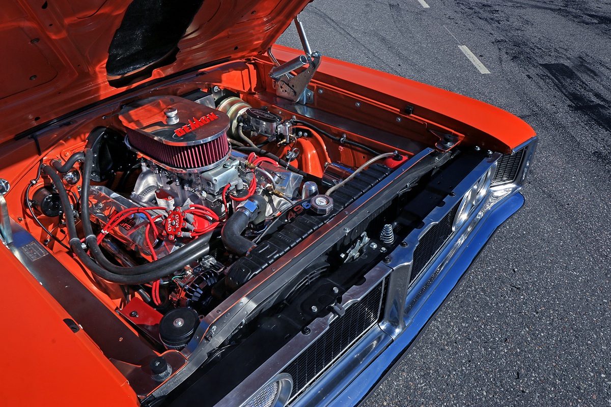 Close-up view of a classic car with its hood open, revealing a clean, high-performance engine with red wiring and shiny metal components. The car’s exterior is bright orange and parked on a paved surface.