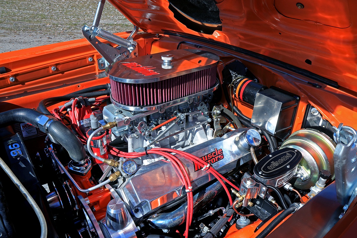Close-up of a high-performance car engine in an orange muscle car, featuring chrome components, red ignition wires, a large air filter, and various mechanical parts under the open hood.