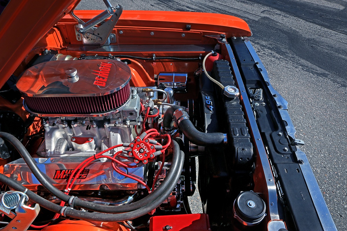 A close-up view of a car’s engine bay with the hood open. The engine is polished with red accents and visible wiring. The car’s exterior is orange and the vehicle is parked on an asphalt surface.