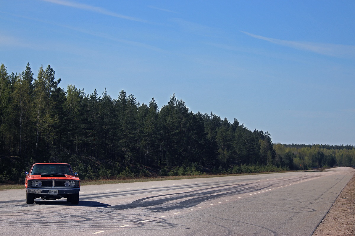 A red vintage muscle car drives on an empty, wide road bordered by tall green trees under a clear blue sky. Tire skid marks are visible on the pavement.