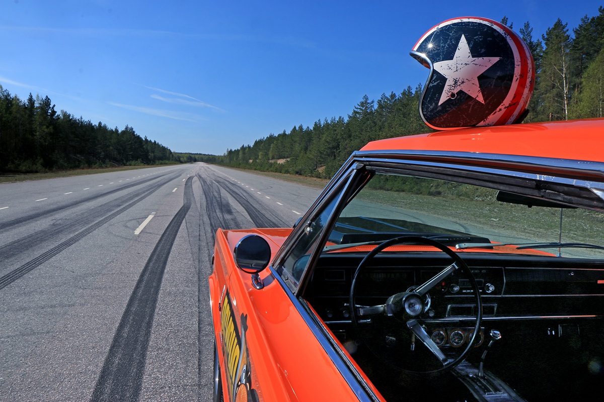 An orange vintage car with a star-painted helmet on its roof is parked on an empty highway. Tire skid marks are visible on the road, surrounded by trees under a clear blue sky.