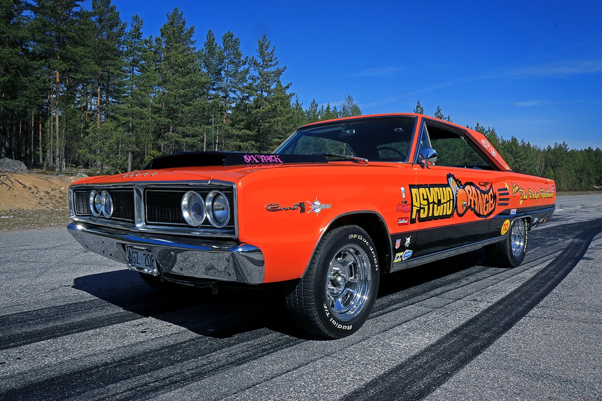 An orange vintage muscle car with “Psycho Garage” graphics and various stickers is parked on a road, surrounded by trees under a blue sky.
