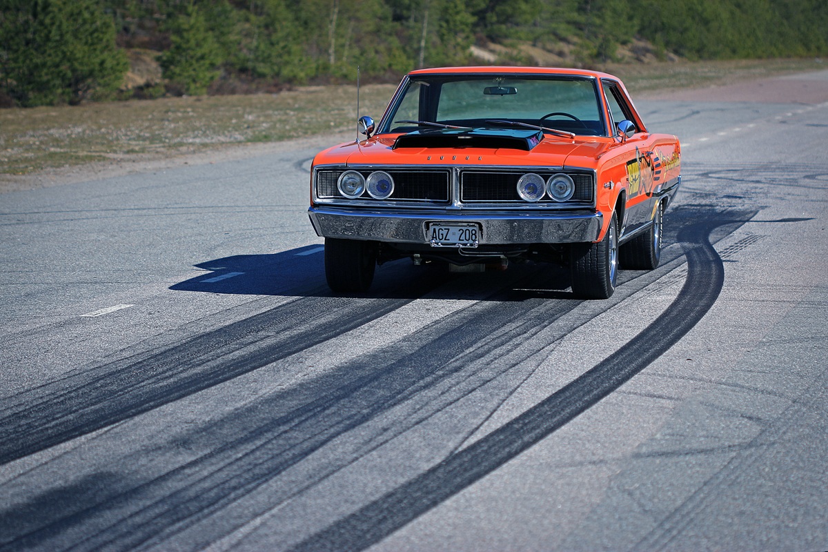 An orange vintage Dodge muscle car drives on a road, leaving dark tire skid marks behind it. The background features trees and a clear sky.