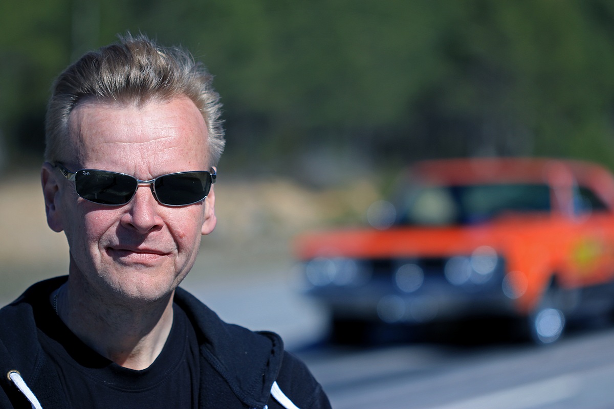 A man with short blond hair wearing sunglasses and a black jacket stands outdoors, smiling slightly. In the blurred background, a red vintage car is parked on a road with trees behind it.