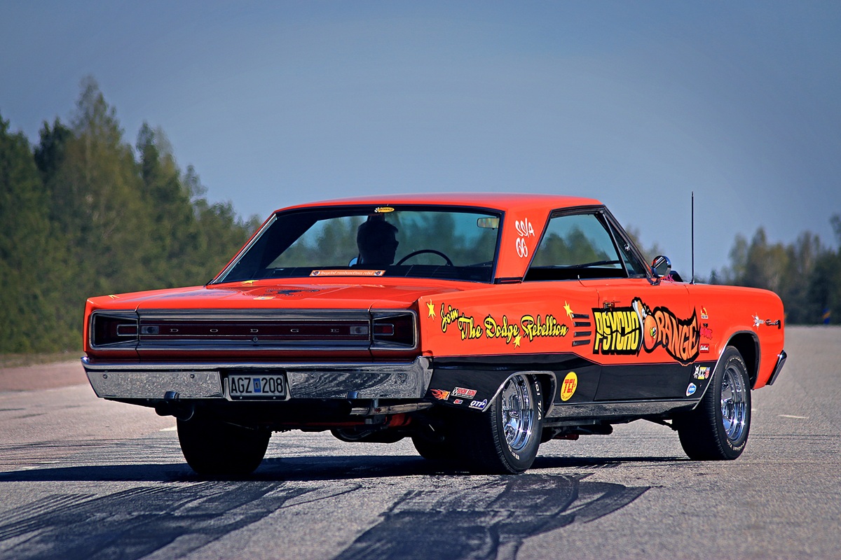 A bright orange vintage muscle car with racing decals and large rear tires is parked on a paved road, with trees and blue sky in the background. The car’s rear is in focus, showing custom artwork and text.