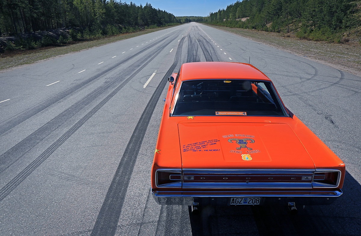 An orange Dodge muscle car parked on an empty, wide road with tire skid marks, surrounded by green trees under a clear blue sky.