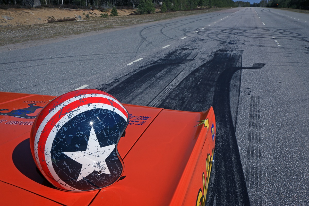 A red, white, and blue helmet with a large white star rests on the hood of an orange car parked on a road with visible tire skid marks and circular burnout patterns. Trees line the road in the background.