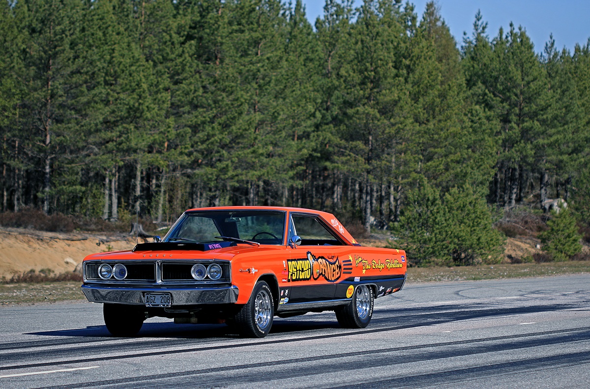 A vintage orange and red muscle car with racing decals drives on a road, surrounded by pine trees and a clear sky in the background.