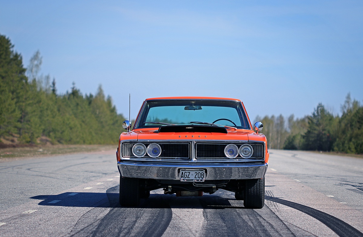 A vintage orange muscle car is parked on an empty road, surrounded by trees and blue sky, viewed from the front with tire marks visible on the pavement.