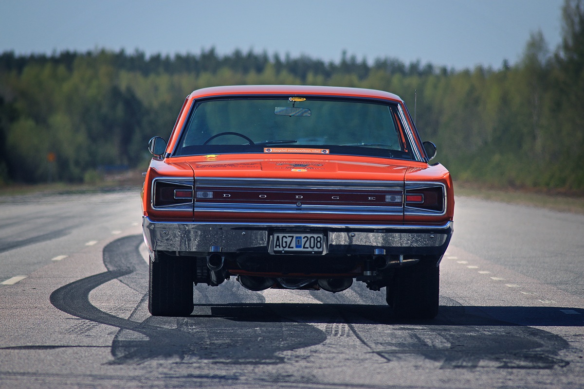Rear view of an orange classic Dodge muscle car on an empty road, with visible tire marks on the pavement and a forest background.