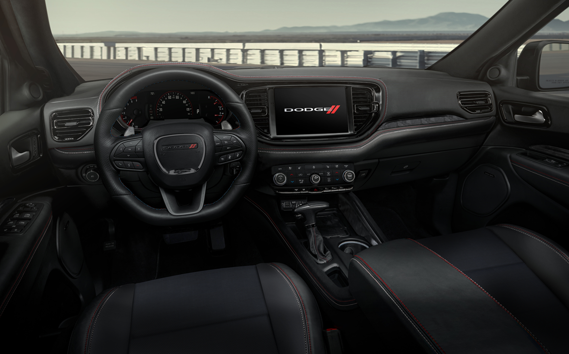 Interior view of a Dodge durango showing the steering wheel, dashboard with digital displays, touchscreen with Dodge logo, and black leather seats with red stitching. A scenic view is visible through the windshield.