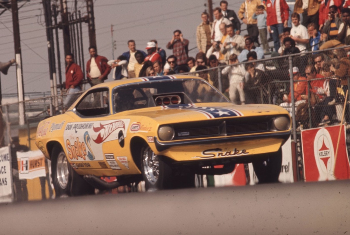 A yellow drag racing car with Snake on the front and sponsor decals speeds down a track with its wheels lifted, while spectators watch from behind a fence.