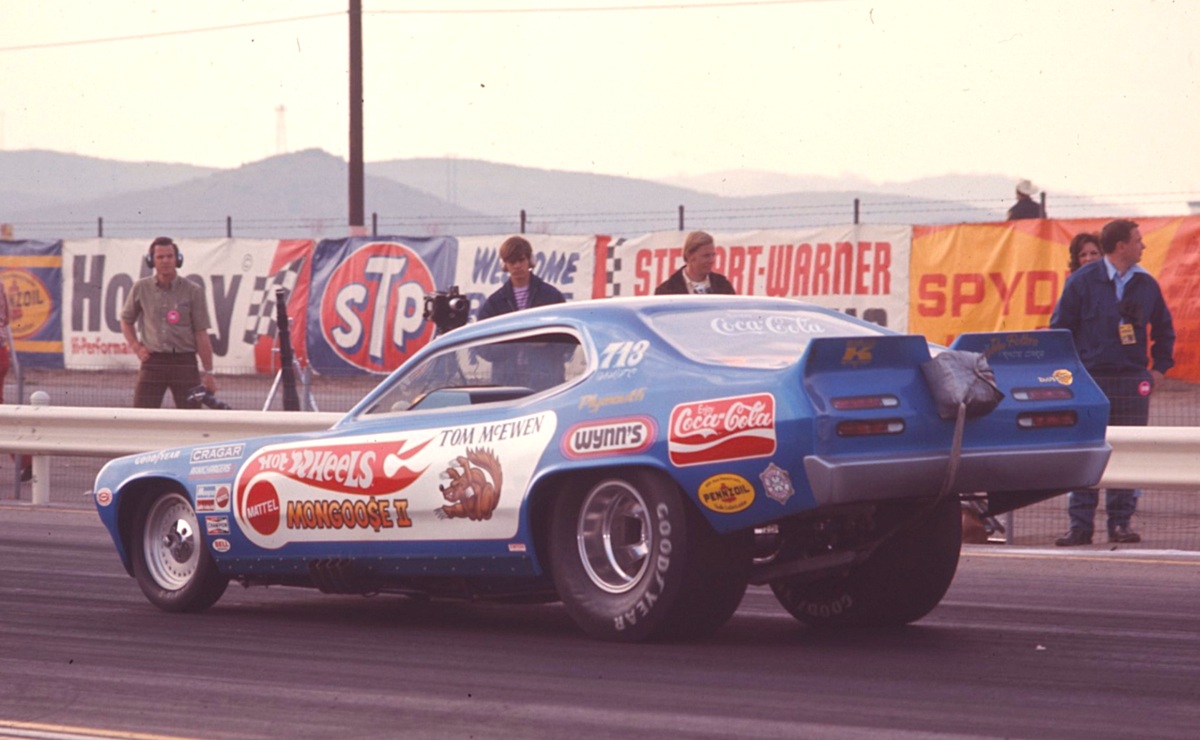 A blue Hot Wheels-sponsored drag racing car, labeled Mongoose II, is at a racetrack starting line with parachutes at the back. Several people and colorful sponsor banners are visible in the background.