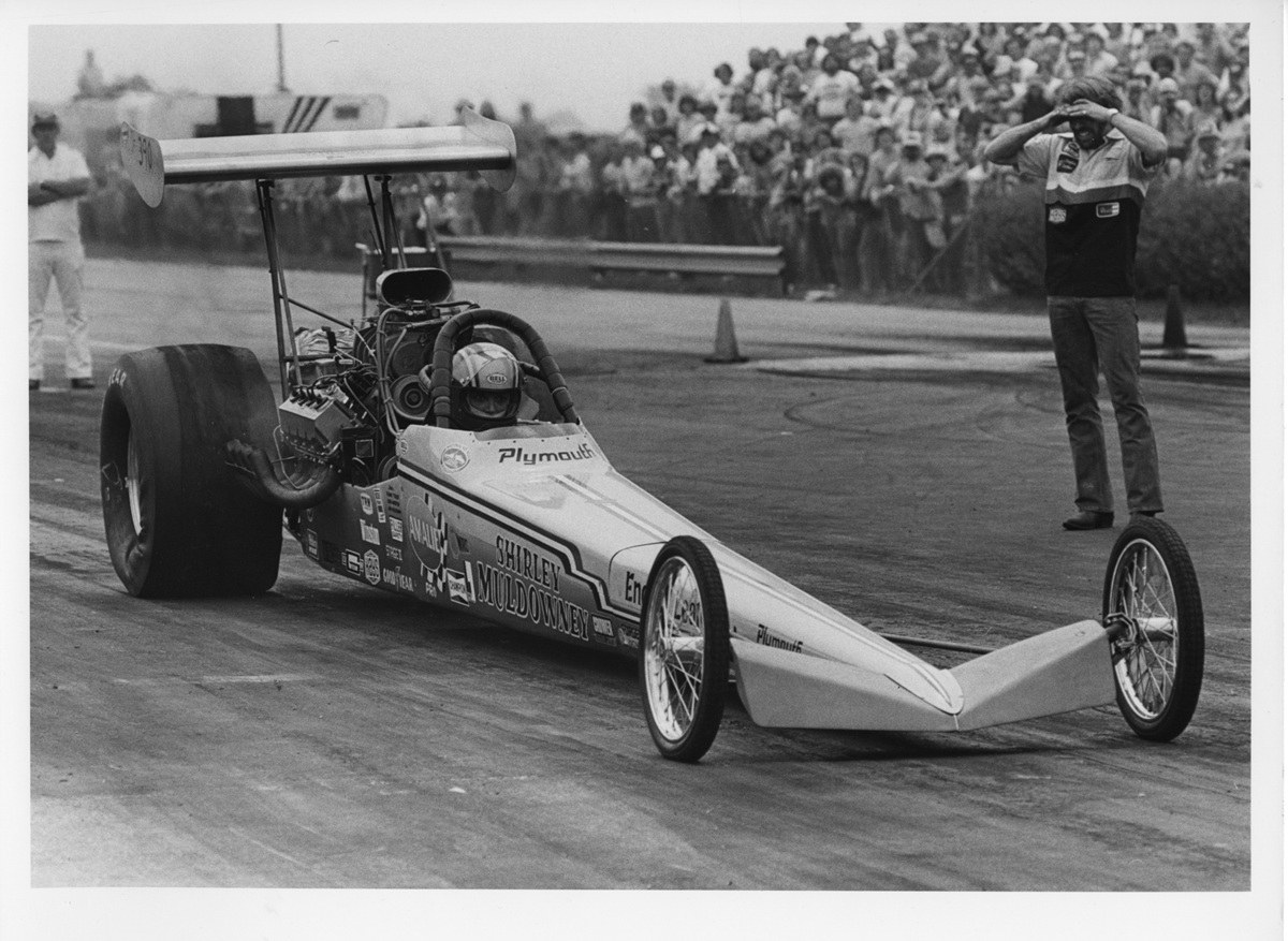 A dragster car with “Plymouth” branding is on a racetrack, preparing to race. A crowd watches in the background while an official stands nearby with hands on his head. The scene appears to be from a past era.