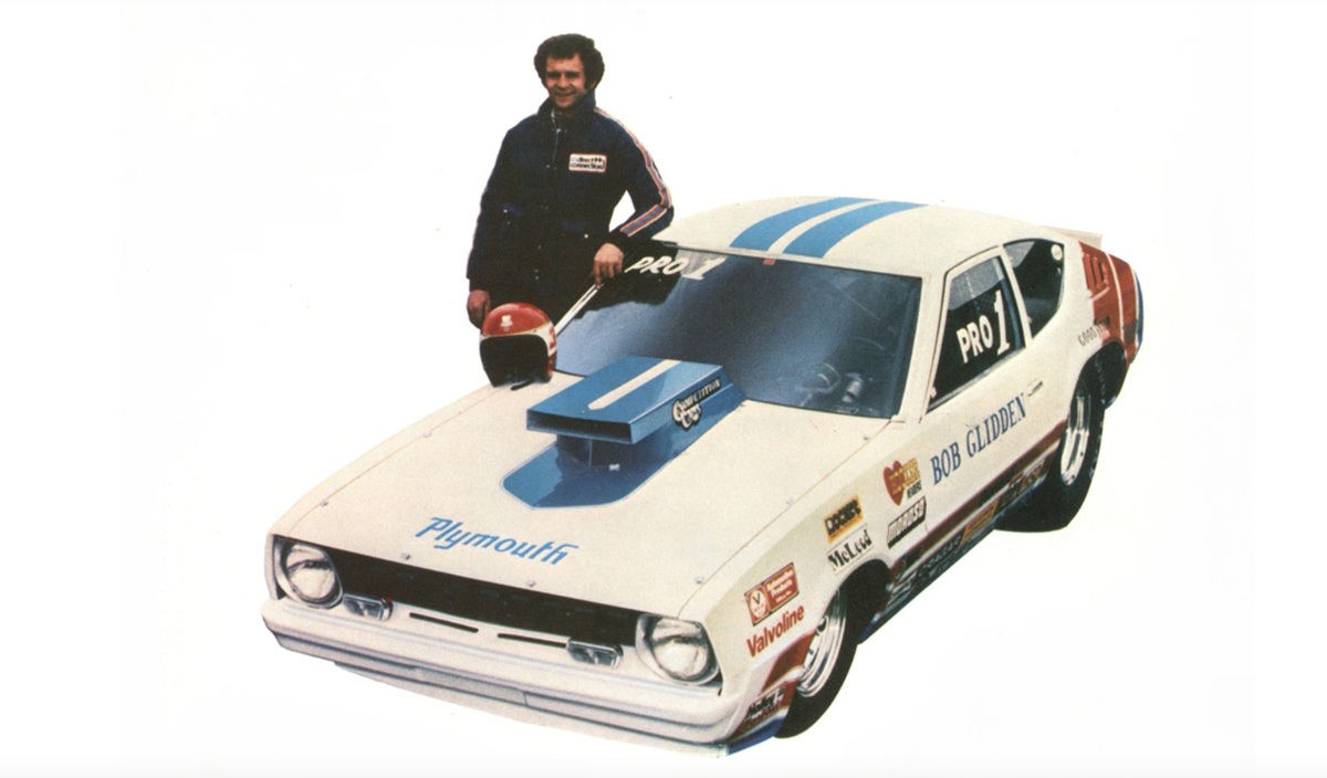 A man in a racing suit stands next to a white Plymouth race car with blue stripes, sponsor decals, and a hood scoop, holding a helmet against a plain white background.