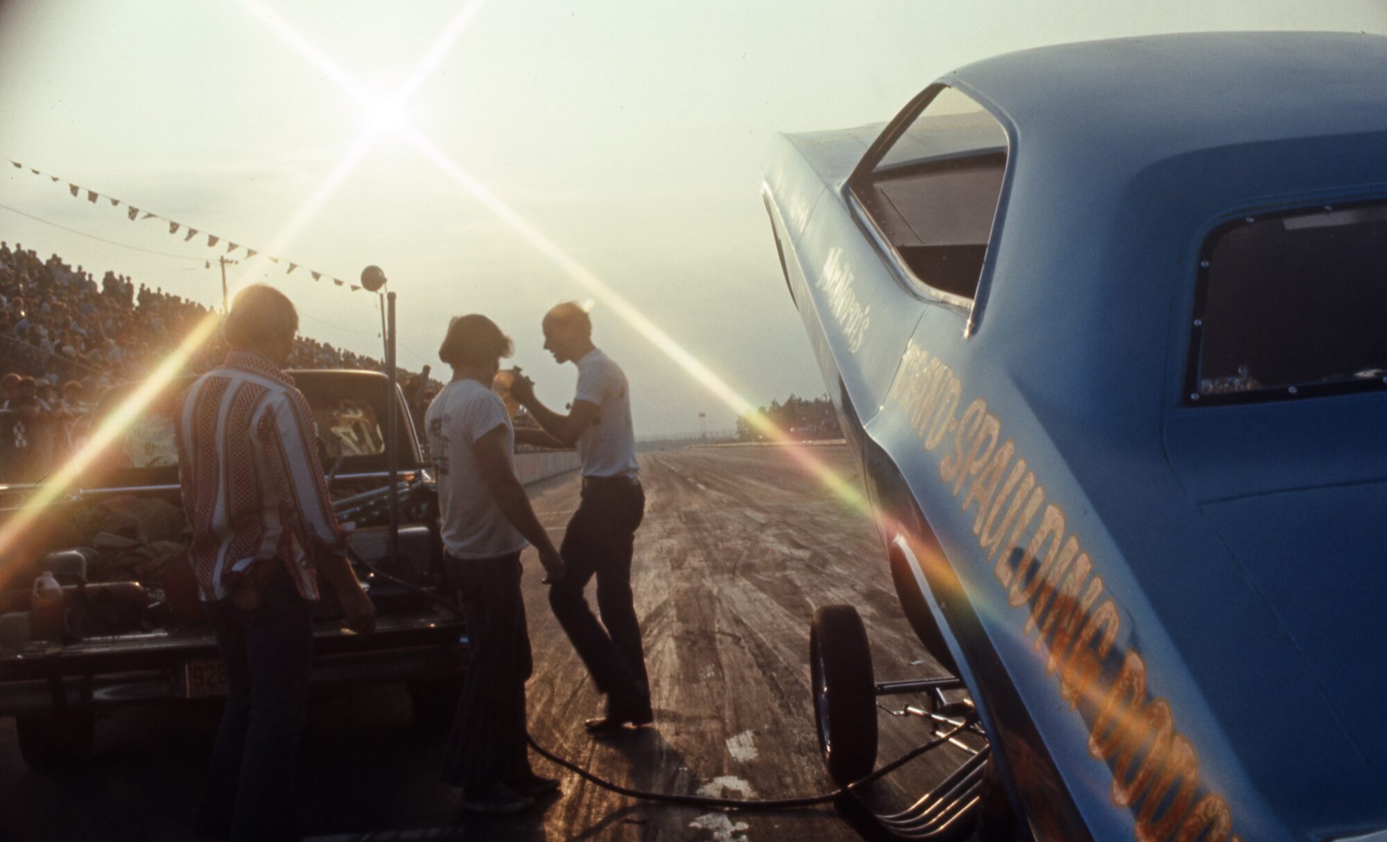 A group of people stand near a blue drag racing car with its body lifted at a sunlit racetrack. The sun shines brightly, casting long shadows, and spectators fill the stands in the background.