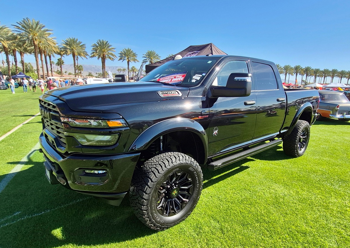 A shiny black lifted pickup truck with large off-road tires is parked on green grass at an outdoor car show, with palm trees and a clear blue sky in the background.
