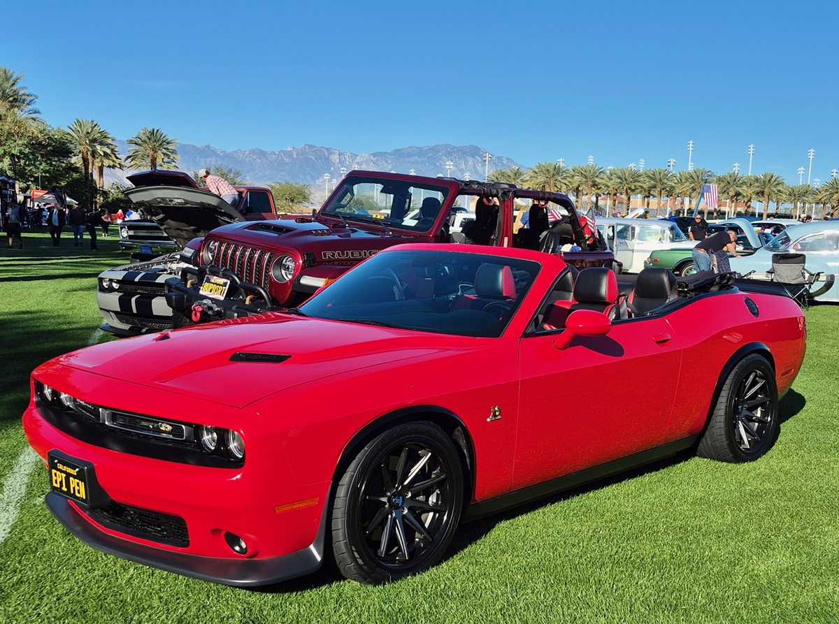 A bright red convertible car is parked on grass at a car show, with its top down. Behind it, there’s a red Jeep with its hood open and palm trees and mountains in the background under a clear blue sky.