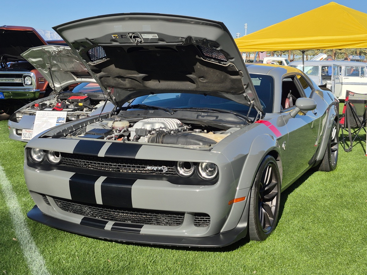 A gray Dodge Challenger with black racing stripes has its hood open, revealing the engine, and is parked on grass at an outdoor car show under a bright yellow canopy.