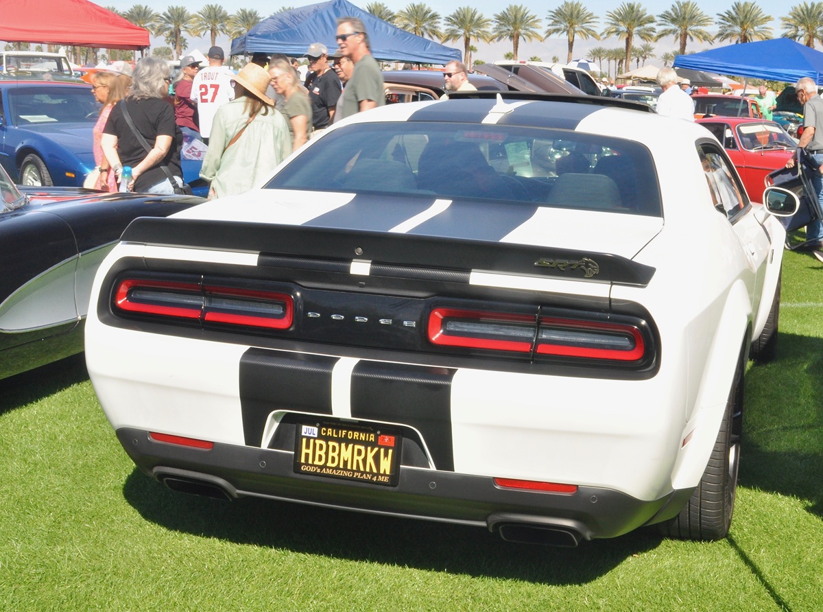 A white Dodge Challenger with black racing stripes is parked on grass at a car show, surrounded by other classic cars and people. The California license plate reads HBMNRKN.