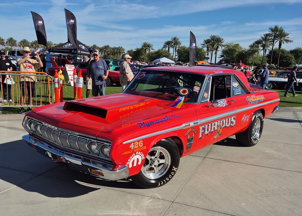 A bright red vintage Plymouth drag racing car Furious is displayed outdoors at a car show, decorated with sponsor stickers, racing numbers, and a winner’s ribbon on the windshield. People and tents are visible in the background.