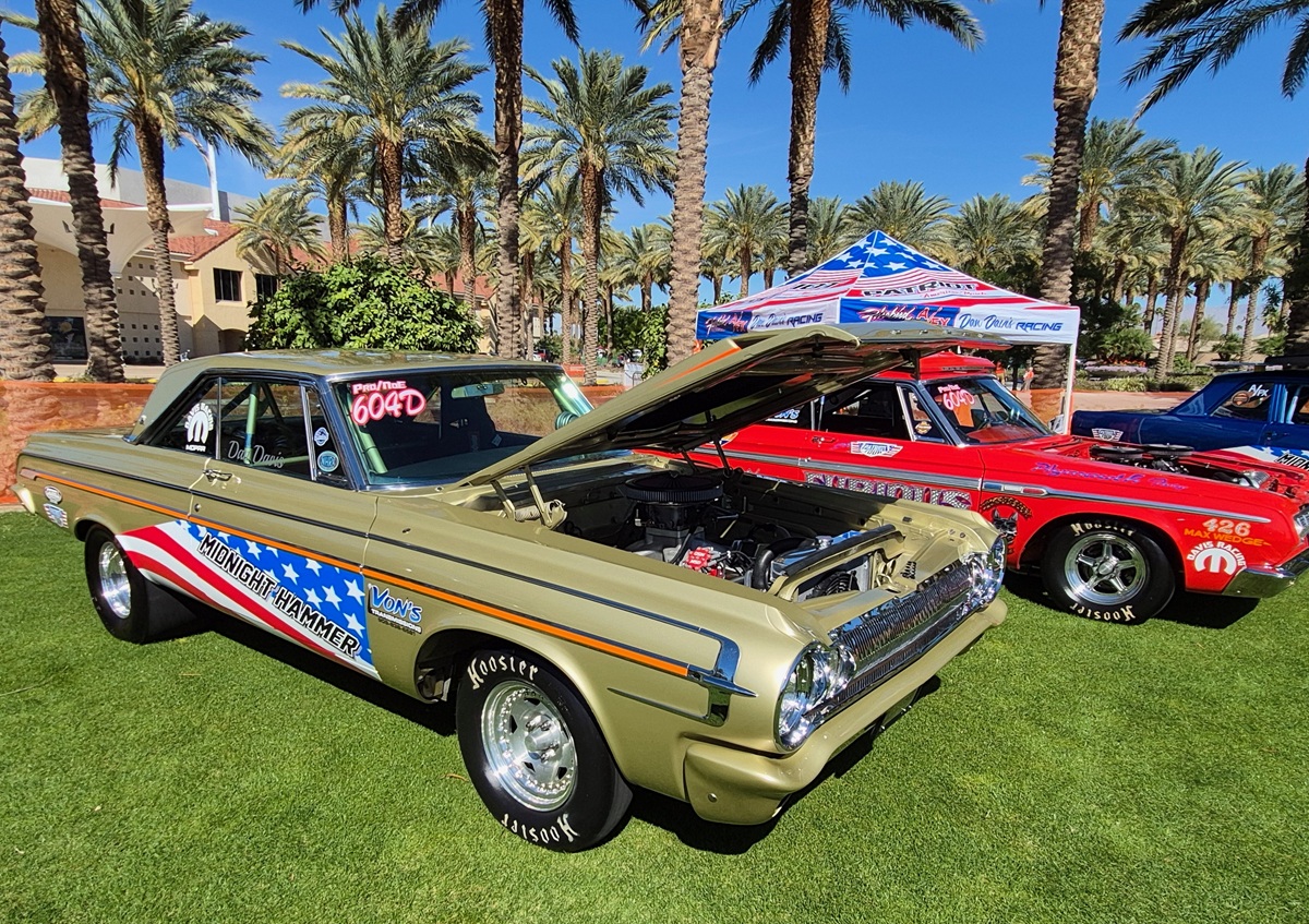 A vintage gold muscle car with its hood open is displayed on grass next to a red race car under palm trees. Both cars feature racing decals and an American flag-themed tent is visible in the background.