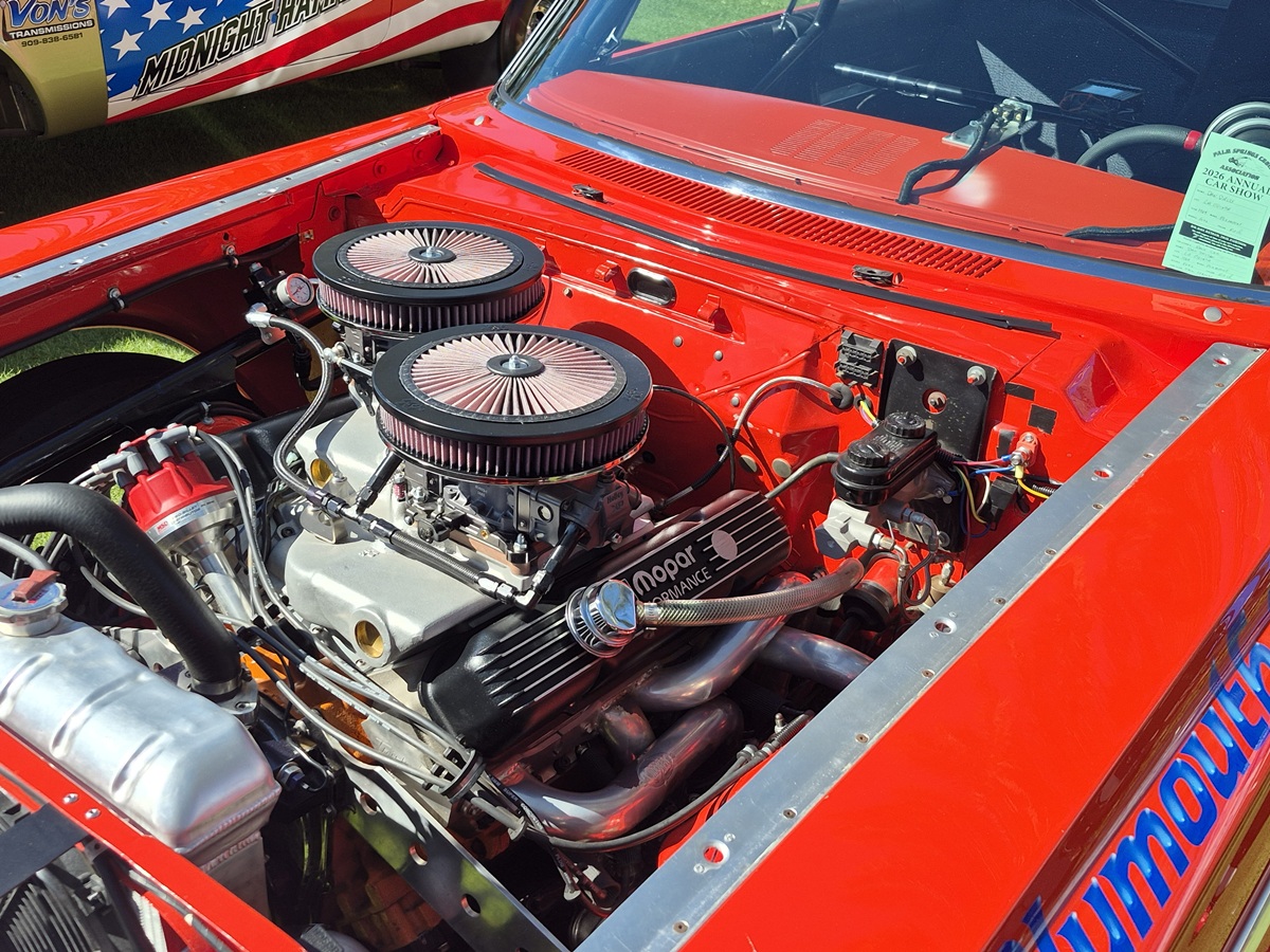 Close-up of a bright red Plymouth car with its hood open, showcasing a powerful Mopar engine with dual air filters and detailed chrome and black components. A car show information sheet is visible on the windshield.