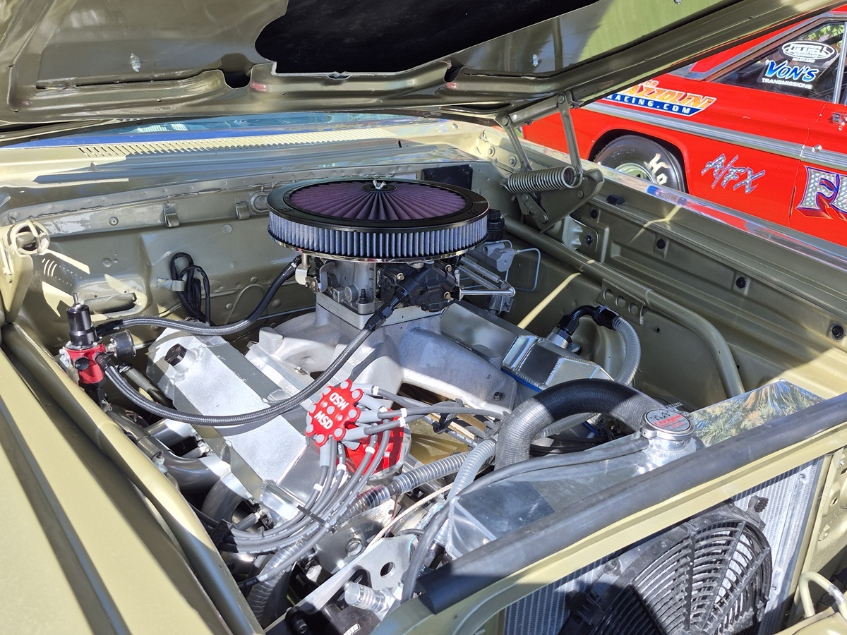 Close-up of a high-performance car engine under an open hood at a car show, with another classic race car featuring bright decals visible in the background.