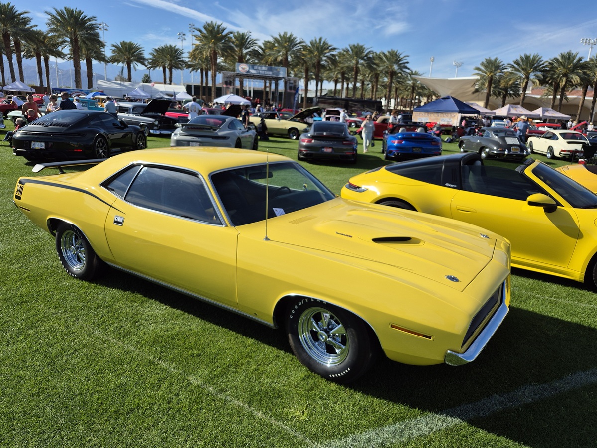 A bright yellow classic muscle car is parked on a grassy field at a car show, surrounded by other colorful vehicles and palm trees under a clear blue sky.