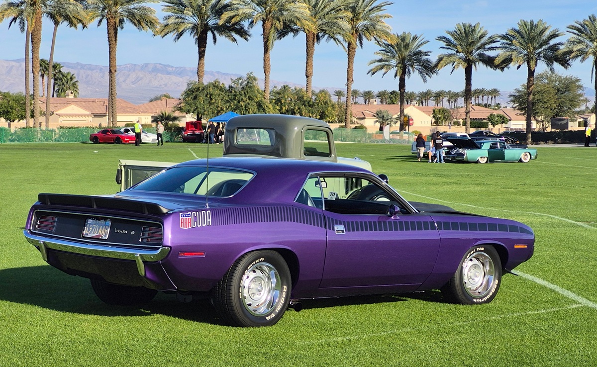 A classic purple Plymouth Barracuda muscle car is parked on a grassy field with palm trees and mountains in the background. Other vintage cars are also visible in the distance under a clear blue sky.