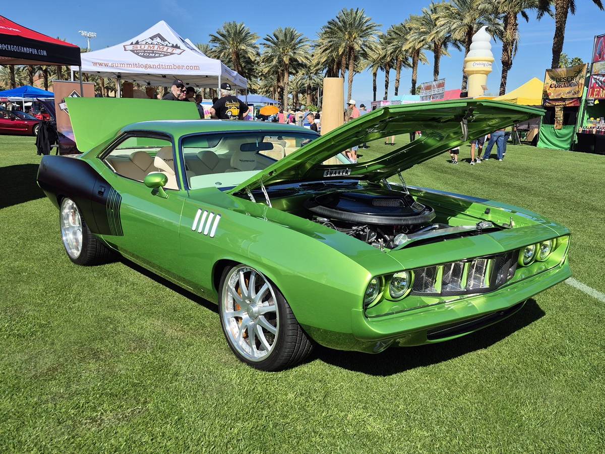 A bright green classic muscle car with its hood open is displayed at an outdoor car show on grass, surrounded by palm trees, people, and tents under a sunny sky.