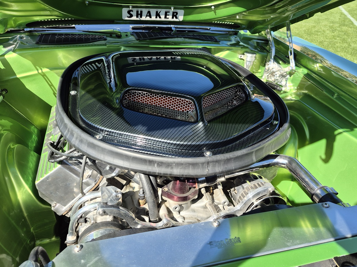 Close-up of a shiny, green classic car engine bay with a prominent black Shaker hood scoop featuring carbon fiber details and mesh vents, reflecting sunlight.