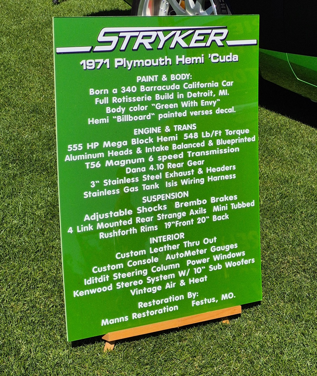A green sign titled STRYKER 1971 Plymouth Hemi Cuda lists detailed specifications of the car’s paint, body, engine, transmission, suspension, and interior, standing on grass beside the vehicle.