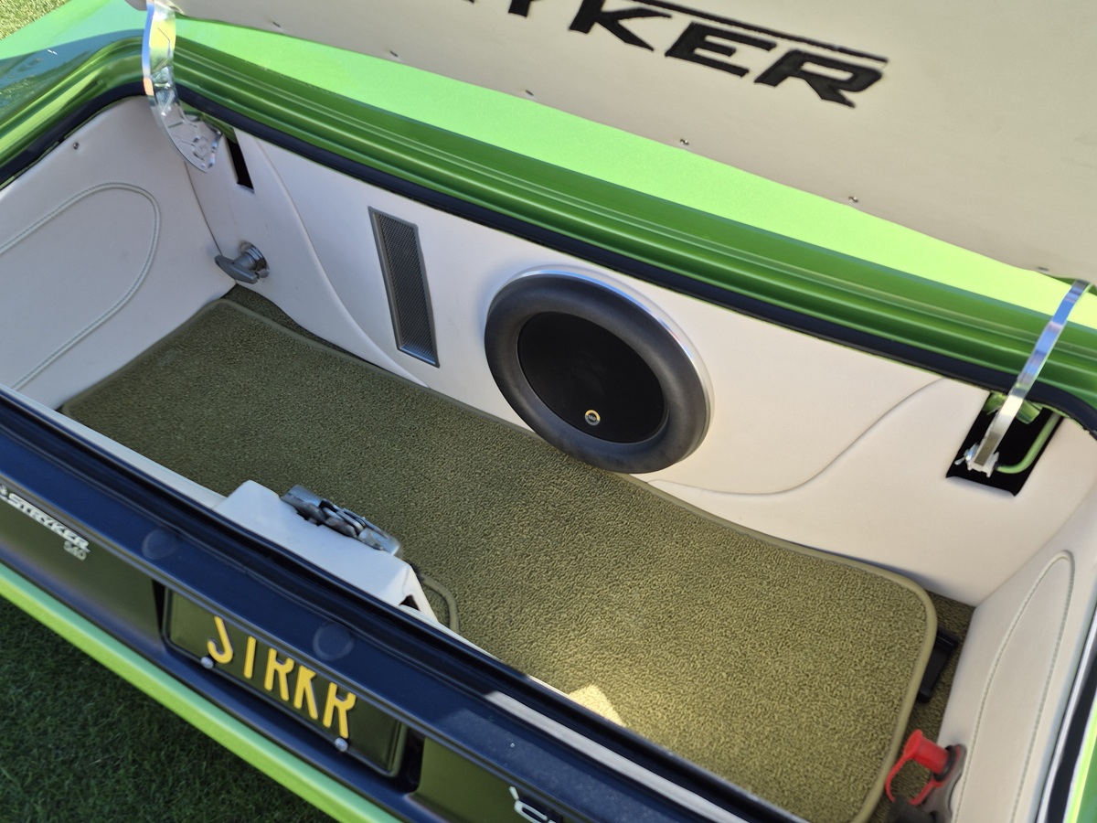 The trunk of a green car with custom beige and white interior, a large black speaker, carpeted flooring, and a personalized license plate reading SIIRK. The trunk lid is open, revealing the setup.