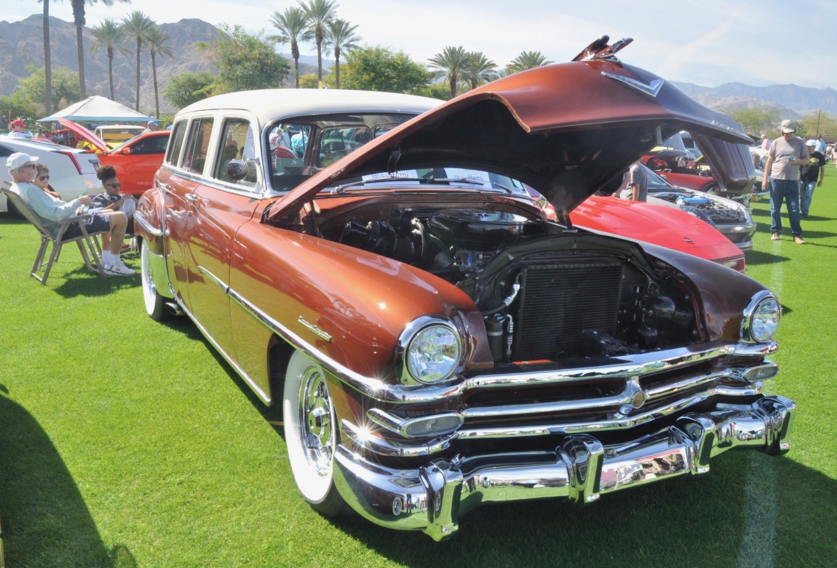 A vintage brown and white station wagon with its hood open is displayed at an outdoor car show on a grassy field, surrounded by people and palm trees under a sunny sky.