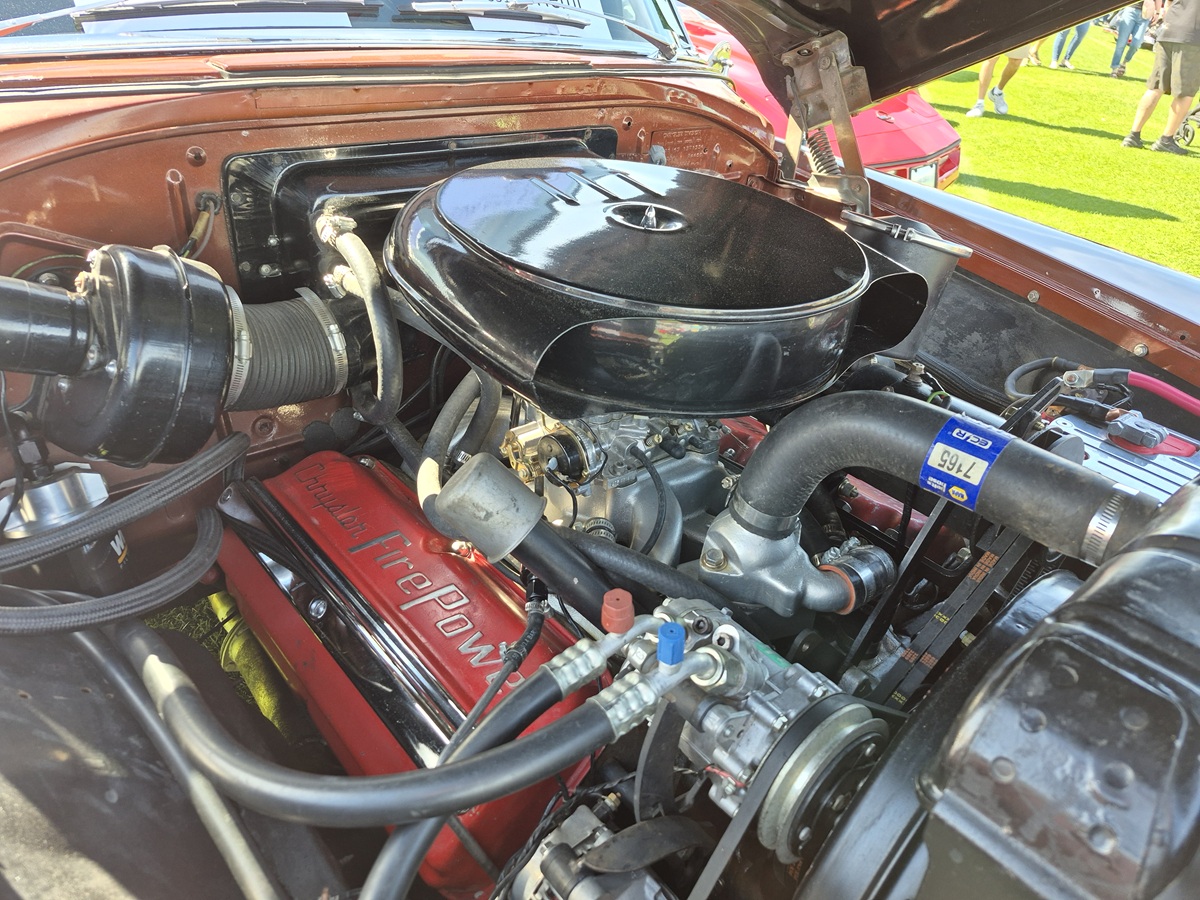 Close-up of a classic car engine under an open hood, featuring a shiny round air filter, red valve covers labeled FirePower, and various hoses and components, with sunlight and grass visible in the background.
