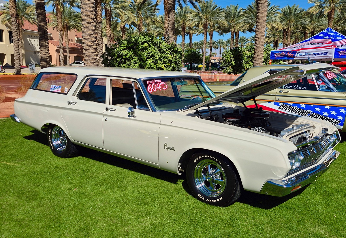 A classic white Plymouth station wagon with an open hood is displayed on grass at a car show, surrounded by palm trees and other vintage cars under a sunny sky.
