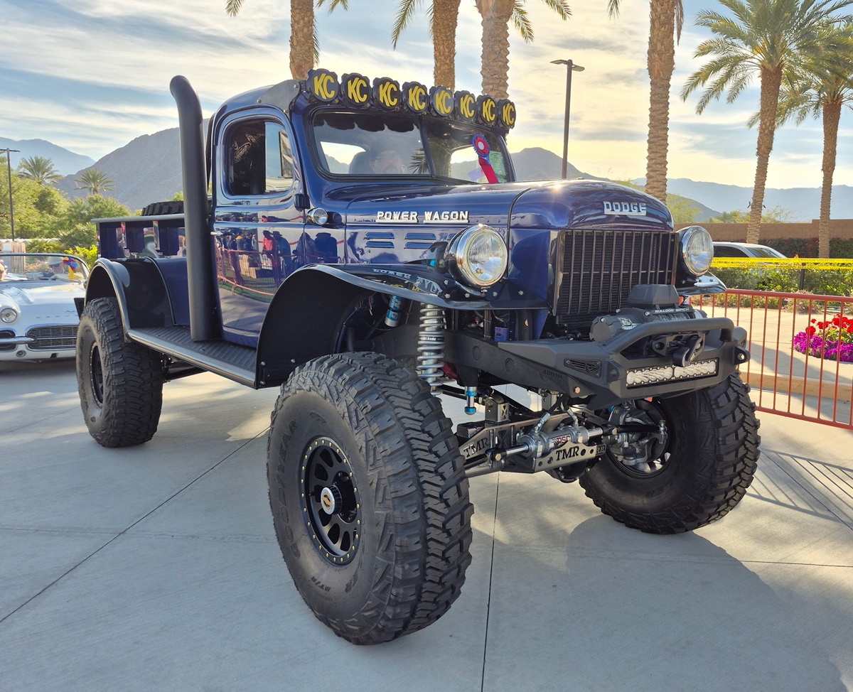 A lifted, restored vintage Dodge Power Wagon truck with large off-road tires is parked outdoors near palm trees. The truck is dark blue and has KC lights mounted on top. Mountains and a classic car are visible in the background.