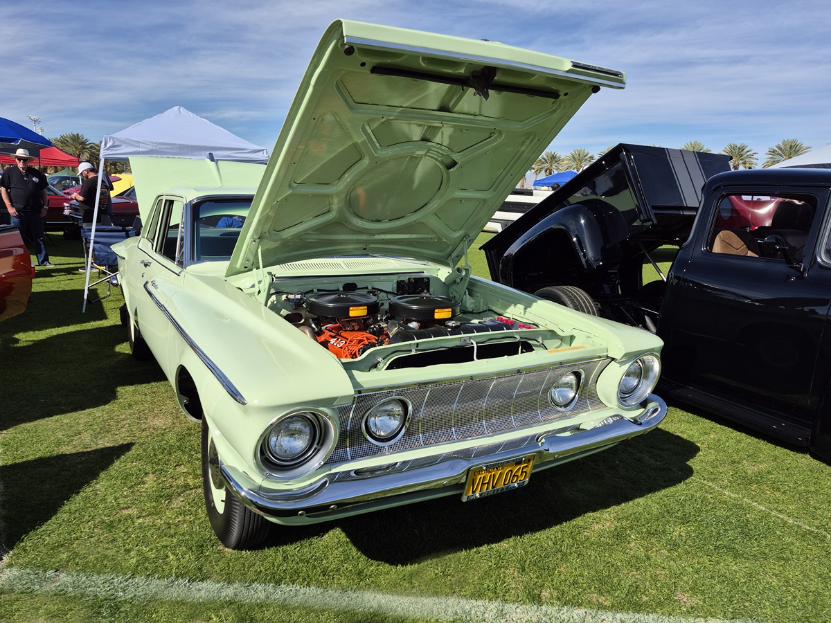 A pale green classic car with its hood open, displaying the engine, is parked on grass at a car show. Other vintage vehicles and tents are visible in the background under a blue sky.