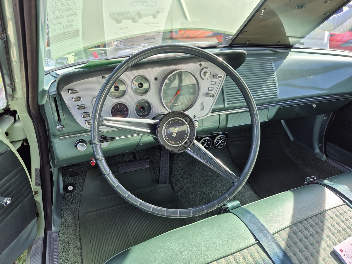 The image shows the interior of a vintage car, focusing on the drivers seat, dashboard, and large steering wheel with a classic design. The dashboard features retro gauges and controls in shades of green.