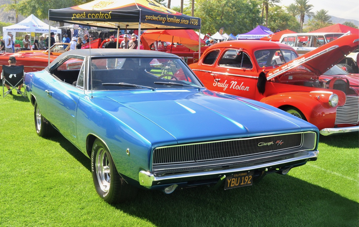 A bright blue vintage Dodge Charger R/T coupe parked on grass at a car show, with a red classic car and event tents in the background.