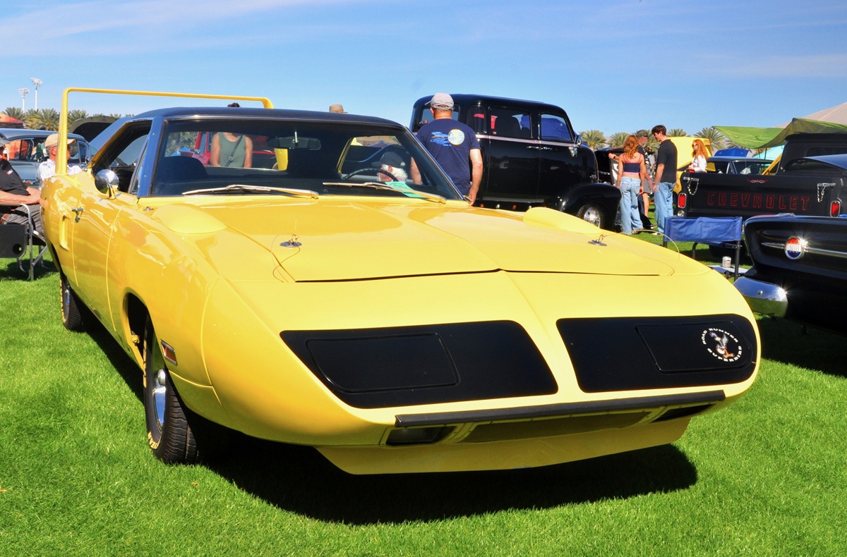 A bright yellow classic muscle car with a prominent rear wing and black accents is parked on grass at an outdoor car show, surrounded by other vintage vehicles and people.
