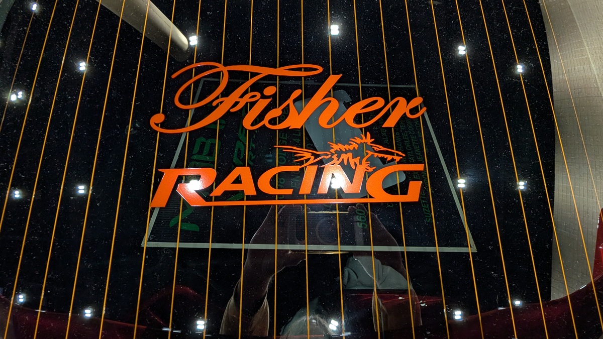 Orange Fisher Racing logo with a stylized fish graphic is seen through a cars rear window, with defroster lines running vertically and bright lights reflecting on the glass.