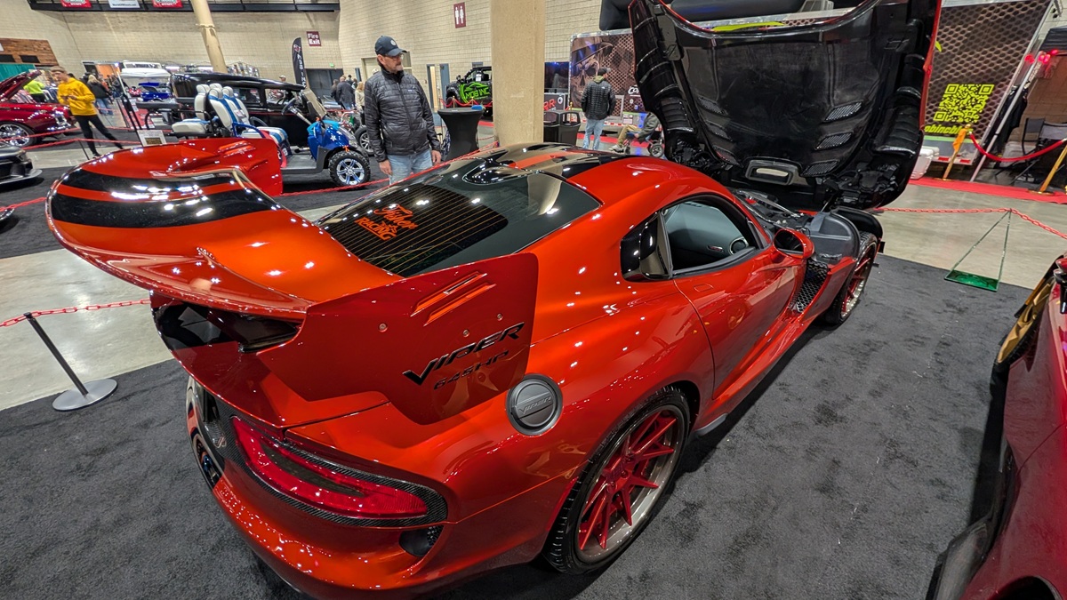 A shiny red Dodge Viper sports car with a large rear wing is displayed at an indoor car show, its hood open. Other custom cars and a few people are visible in the background.