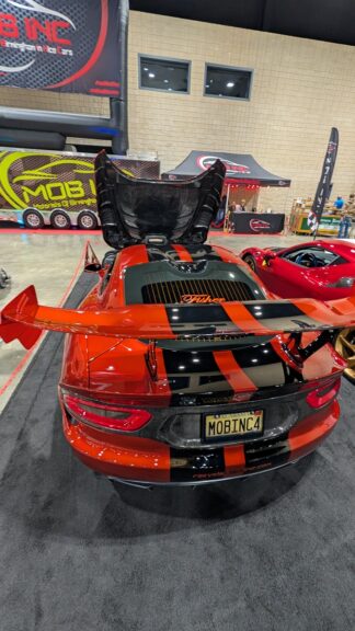 A red and black Dodge Viper with its rear hatch open is displayed indoors at a car show. The car has a large rear wing and a custom license plate reading “MOBINC4.” Other cars and banners are visible in the background.