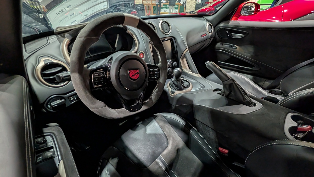 A close-up view of the interior of a sports car, showing a black suede steering wheel with a red snake emblem, manual gearshift, touchscreen, and black leather seats with white stitching.