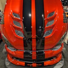 A red and black sports car with its hood open, featuring prominent air vents and bold racing stripes, displayed indoors at an auto show.