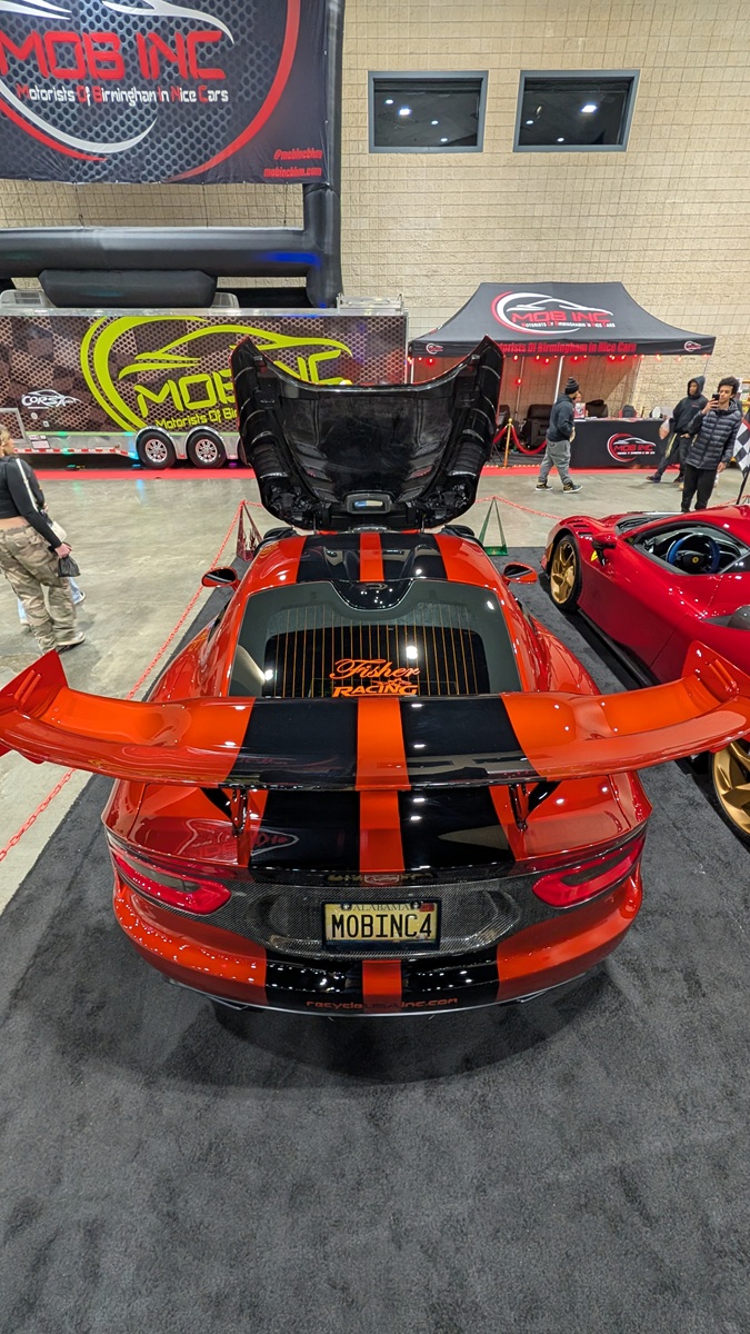Bright orange and black sports car displayed indoors with its hood open, large rear wing, and custom license plate reading MOBINC4. People and other cars are visible in the background at the car show.