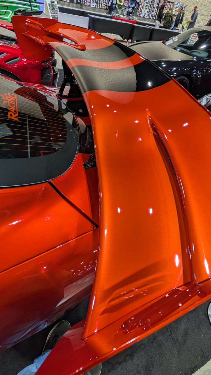 Close-up view of a shiny, metallic orange sports car with a large rear spoiler, black accents, and reflections from indoor lighting at a car show. Other vehicles and display tables are visible in the background.