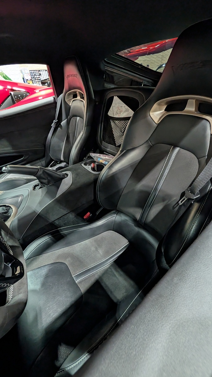 Interior view of a sports car featuring two black and gray leather seats with sporty designs, a center console, and visible seatbelts. The car door and parts of a red car outside are also visible.